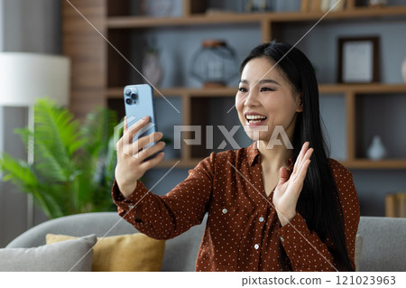Asian woman smiling during video call, sitting on couch with smartphone in hand. Wearing polka dot shirt, expressing happiness and friendliness at home. Modern setting with decor. Asian woman smiling during video call, sitting on couch with smartphone in hand. Wearing polka dot shirt, expressing happiness and friendliness at home. Modern setting with decor. 121023963