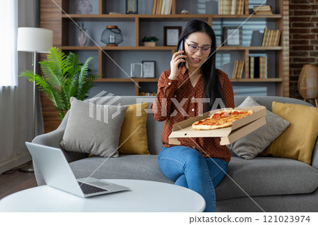 Asian woman enjoying phone conversation while holding pizza box at home, creating relaxed atmosphere. Laptop on table suggests remote work. Casual yet productive home setting. 121023974