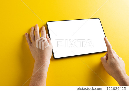 Hands of caucasian woman using tablet with blank screen and copy space on yellow background 121024425