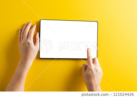 Hands of caucasian woman using tablet with blank screen and copy space on yellow background 121024426
