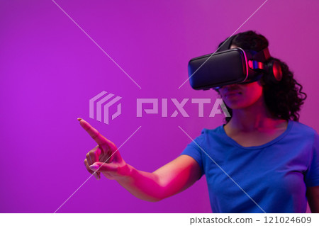 Biracial woman using vr headset and pointing on neon pink to purple background 121024609