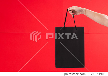 Hand of caucasian woman holding black gift bag with copy space on red background Hand of caucasian woman holding black gift bag with copy space on red background 121024908
