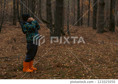 A lost child in orange boots and a warm jacket screams for help in a deserted autumn forest with his hands clasped around his mouth. The child decided to take a walk alone in the forest and got lost. 121025050