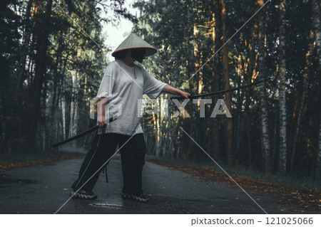 A martial artist in a conical Asian hat practices fighting techniques with a katana sword on a road in the forest. Katana training in the forest against the backdrop of autumn trees. A martial artist in a conical Asian hat practices fighting techniques with a katana sword on a road in the forest. Katana training in the forest against the backdrop of autumn trees. 121025066