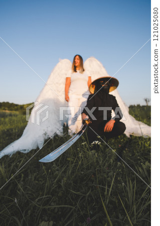 Angel and samurai with katana against clear sky. Katana sword in hands of ninja in suit, blurred angel with big white wings standing in background, green meadow, sunset, clear sky. 121025080