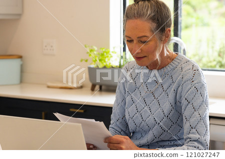 Mature woman reviewing documents at home, focused and thoughtful in kitchen Mature woman reviewing documents at home, focused and thoughtful in kitchen 121027247