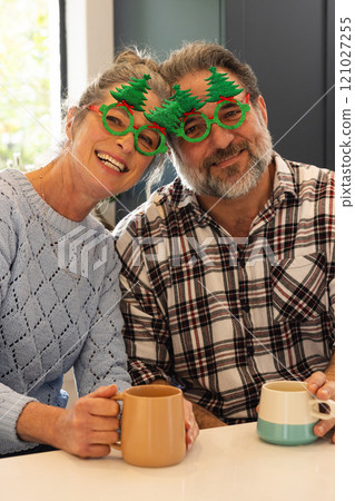 Christmas time, Mature couple wearing festive glasses enjoying coffee and smiling at home 121027255