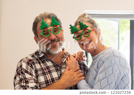 Mature couple wearing festive glasses smiling joyfully at home during Christmas Mature couple wearing festive glasses smiling joyfully at home during Christmas 121027256