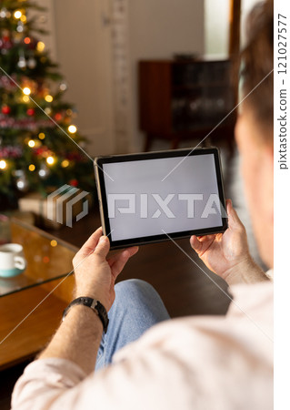 senior man using tablet at home near Christmas tree, enjoying holiday season, copy space 121027577
