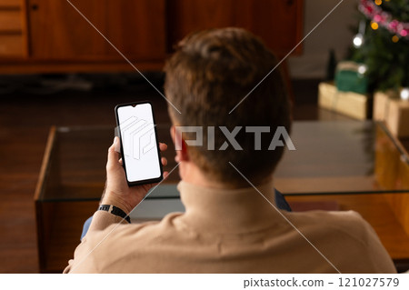 senior man using smartphone at home near Christmas tree, relaxing on couch, copy space 121027579