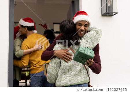 On doorstep, multiracial friends exchanging gifts and hugging at christmas party, wearing santa hats 121027655
