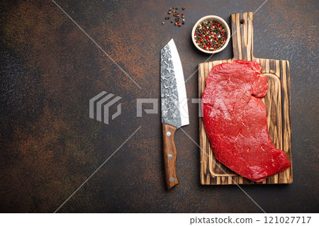 Raw uncooked top round beef steak on wooden cutting board with big kitchen knife and pepper on dark brown rustic stone background top view, cooking meat steak with copy space 121027717