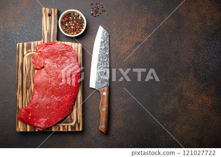 Raw uncooked top round beef steak on wooden cutting board with big kitchen knife and pepper on dark brown rustic stone background top view, cooking meat steak with copy space 121027722