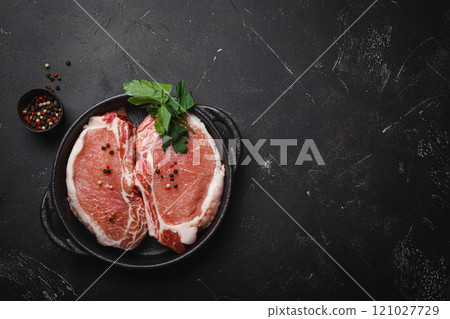 Cut raw meat pork steaks with seasonings in black cast iron pan, dark rustic stone background top view, ready for roasting. Pork loin chops cooking, space for text 121027729