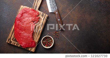 Raw uncooked top round beef steak on wooden cutting board with big kitchen knife and pepper on dark brown rustic stone background top view, cooking meat steak with copy space 121027730