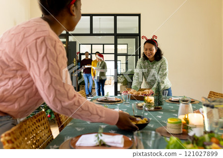 Preparing festive dinner table, multiracial friends wearing christmas accessories,at home 121027899