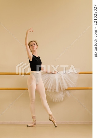 Ballet dancer gracefully raising arm while posing near barre 121028027