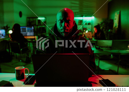 Medium close up of African American brutal hacker programming laptop in room with other hackers in background Medium close up of African American brutal hacker programming laptop in room with other hackers in background 121028111