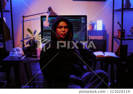 Portrait of female hacker dressed in black hoodie smiling lightly, table with computer, personal stuff and white mask behind her in dimly light room Portrait of female hacker dressed in black hoodie smiling lightly, table with computer, personal stuff and white mask behind her in dimly light room 121028119