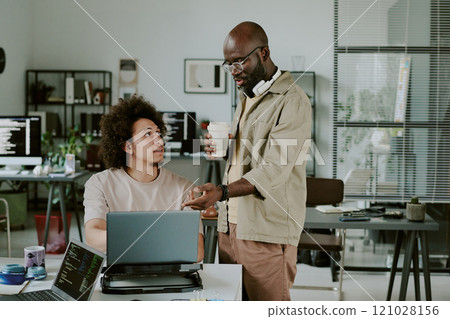 African American IT specialist with cup of fresh brewed coffee helping his colleague with coding African American IT specialist with cup of fresh brewed coffee helping his colleague with coding 121028156