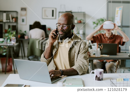 Medium close up of African American office worker working on laptop while speaking on smartphone 121028179
