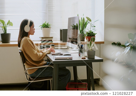 Side view shot of brunette young adult woman listening to music while coding in biophilic office during workday 121028227