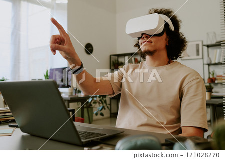 Low angle of young adult man sitting at desk with laptop and gesticulating while using VR goggles Low angle of young adult man sitting at desk with laptop and gesticulating while using VR goggles 121028270