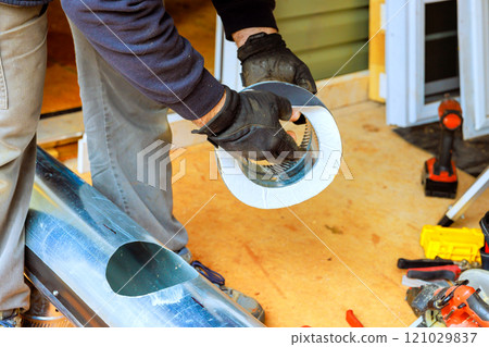 Worker fitting metal heating duct into place during home renovation, using tools materials scattered around. Worker fitting metal heating duct into place during home renovation, using tools materials scattered around. 121029837