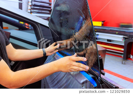 Technician worker applies tint film to car window in well lit automotive workspace, ensuring perfect fit. 121029850