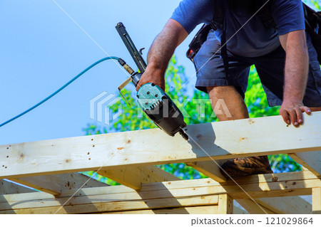 Construction worker fastens wooden beams while standing on roof, utilizing air power nail gun tool during construction at work area 121029864