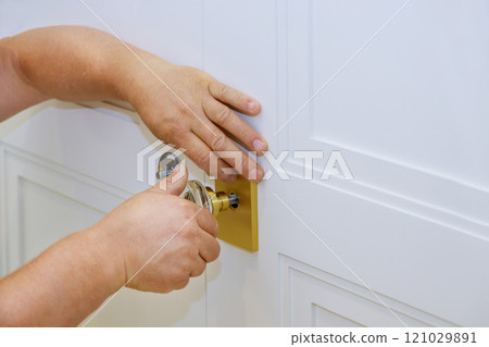 Carpenter is carefully installing door lock on white panel door, during home improvement project. 121029891