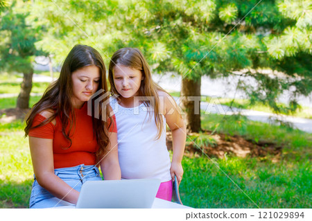 Young girls enjoy learning moment outside on sunny day, focusing intently on laptop they are surrounded by lush greenery enhancing their study setting. Young girls enjoy learning moment outside on sunny day, focusing intently on laptop they are surrounded by lush greenery enhancing their study setting. 121029894