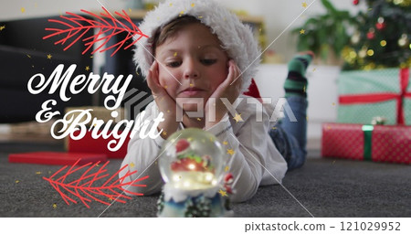 Image of merry and bright over caucasian boy in santa hat with snow ball lying on floor 121029952