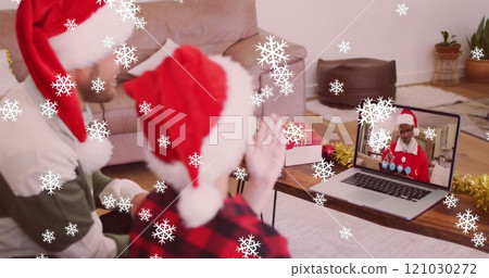 Image of snow falling over smiling father and son in santa hats on laptop video call with family 121030272