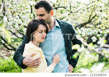 husband and a pregnant wife hug in a spring garden surrounded by blossoming apple trees 121030659