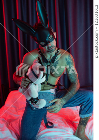 Sexy guy in bdsm rabbit mask and leather belt on a bed in neon light waiting for sex with a mistress with a toy 121031012