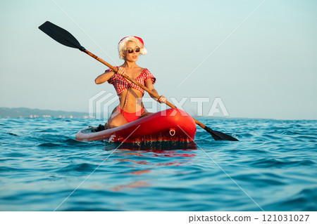Sexy girl in a Santa Claus hat floats on the sea on a Sup Board in a red bikini celebrating Christmas at a resort in a hot country 121031027