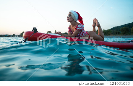 Sexy girl in a Santa Claus hat floats on the sea on a Sup Board in a red bikini celebrating Christmas at a resort in a hot country 121031033
