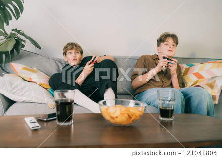 Two teenager boys are focused on their video game consoles while sitting on couch at home. Brothers Gamers enjoying Playing Video Games indoors. Siblings Spending time at home with snacks and drinks. 121031803