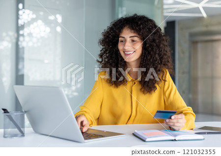Smiling Latin American businesswoman using laptop and holding credit card. Engaged in online transaction or educational task. Represents modern business environment and online work. Smiling Latin American businesswoman using laptop and holding credit card. Engaged in online transaction or educational task. Represents modern business environment and online work. 121032355