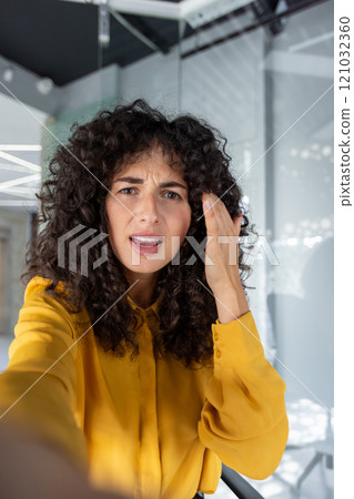 Curly-haired woman making video call using phone, expressing concern or confusion in modern office setting. Bright yellow shirt adds contrast to scene. Emphasizes emotions during call. 121032360
