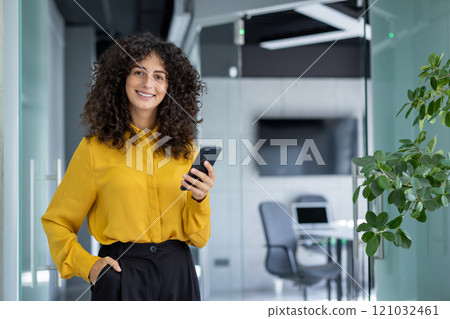 Businesswoman smiling confidently in modern office, holding smartphone. Bright workspace features glass walls and green plant. Woman's attire includes stylish yellow blouse and black pants. 121032461