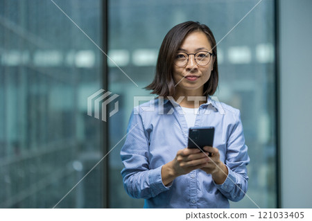 An Asian businesswoman in glasses uses her smartphone confidently in a modern, professional environment. Her poised demeanor conveys a sense of focus and competence. 121033405