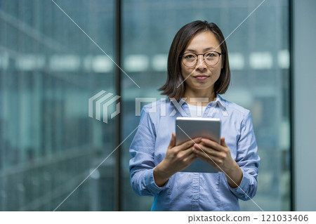 Asian woman confidently holds a tablet in a modern office. She appears professional, wearing glasses and a blue shirt, symbolizing success and technology in a business setting. 121033406