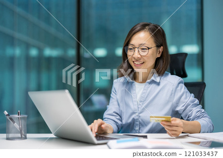 Asian businesswoman using laptop, holding credit card. Smiling, engaged in online shopping or electronic payment. Professional setting emphasizes modern business, finance, technology. Asian businesswoman using laptop, holding credit card. Smiling, engaged in online shopping or electronic payment. Professional setting emphasizes modern business, finance, technology. 121033437