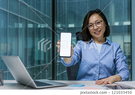 Young businesswoman inside office at workplace showing white phone screen to camera, Asian woman smiling happily advertising mobile online financial app. 121033450