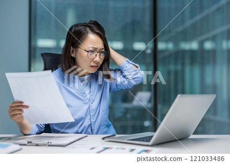 An Asian businesswoman appears frustrated looking at her laptop and holding documents. The scene conveys stress or confusion, possibly due to work-related challenges or deadlines in office setting. 121033486