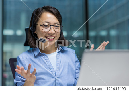 Asian woman wearing a headset is actively participating in a virtual meeting. She is gesturing with her hands and smiling, reflecting communication and engagement in a professional environment. Asian woman wearing a headset is actively participating in a virtual meeting. She is gesturing with her hands and smiling, reflecting communication and engagement in a professional environment. 121033493