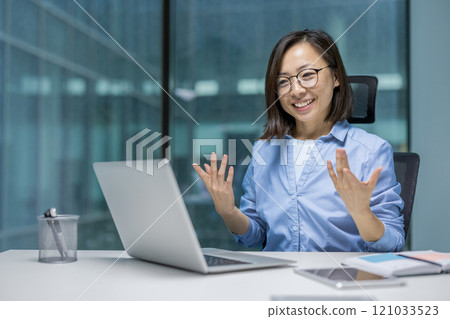 Asian businesswoman smiles conversing in a video call using her laptop. She appears engaged and professional, suggesting a successful online meeting environment within a modern office setting. Asian businesswoman smiles conversing in a video call using her laptop. She appears engaged and professional, suggesting a successful online meeting environment within a modern office setting. 121033523
