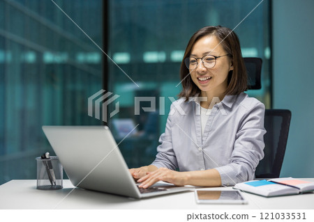 Asian businesswoman sits at desk, smiling while working on laptop. Professional setting reflects productivity and focus. Includes laptop, phone, documents highlighting modern office environment. 121033531